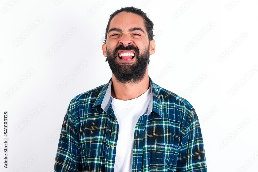 young bearded hispanic man wearing plaid shirt over white background with happy and funny face smiling and showing tongue. young bearded hispanic man wearing plaid shirt over white background with happy and funny face smiling and showing tongue.