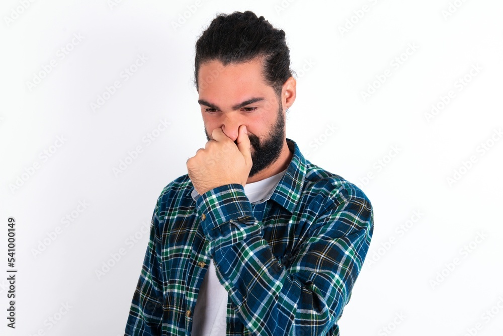 young bearded hispanic man wearing plaid shirt over white background, holding his nose because of a bad smell. young bearded hispanic man wearing plaid shirt over white background, holding his nose because of a bad smell.