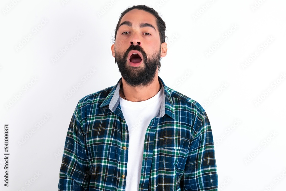 Emotional attractive young bearded hispanic man wearing plaid shirt over white with opened mouth expresses great surprise and fright, stares at camera. Unexpected shocking news and human reaction. Emotional attractive young bearded hispanic man wearing plaid shirt over white with opened mouth expresses great surprise and fright, stares at camera. Unexpected shocking news and human reaction.