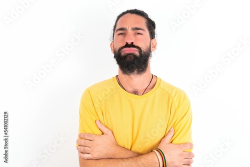 Gloomy dissatisfied young bearded hispanic man wearing yellow T-shirt over white background looks with miserable expression at camera from under forehead, makes unhappy grimace