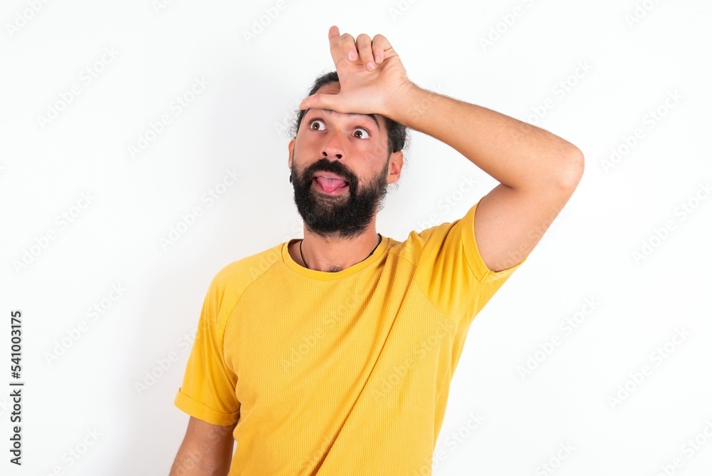 young bearded hispanic man wearing yellow T-shirt over white background gestures with finger on forehead makes loser gesture makes fun of people shows tongue young bearded hispanic man wearing yellow T-shirt over white background gestures with finger on forehead makes loser gesture makes fun of people shows tongue