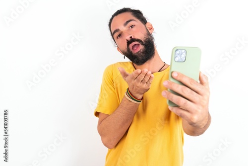 young bearded hispanic man wearing yellow T-shirt over white background blows air kiss at camera of smartphone and takes selfie, sends mwah via online call.