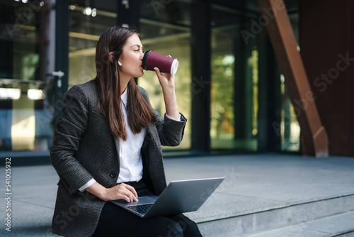 Young woman working online on laptop computer sitting outside the office building wearing wireless headphones enjoying a takeaway coffee. Freelancer working