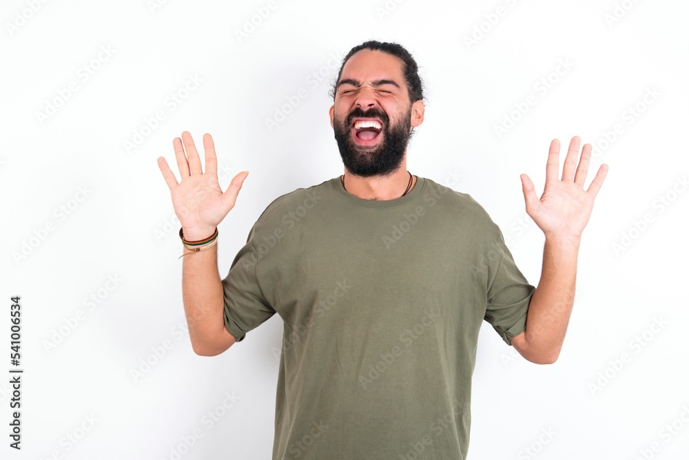 Naklejka premium Emotive young bearded hispanic man wearing green T-shirt over white background laughs loudly, hears funny joke or story, raises palms with satisfaction, being overjoyed amused by friend