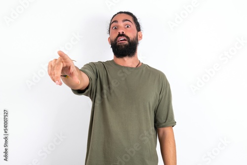 young bearded hispanic man wearing green T-shirt over white background Pointing with finger surprised ahead, open mouth amazed expression, something on the front.