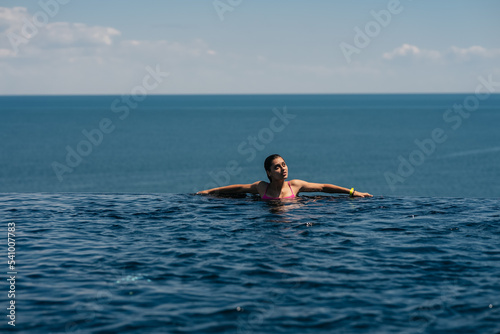 Happy woman in swimsuit swimming in infinity pool against seafront.