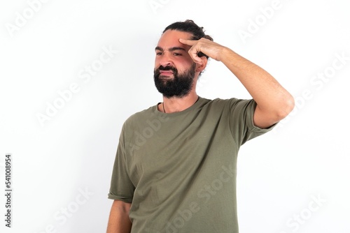 young bearded hispanic man wearing green t-shirt over white background pointing unhappy at pimple on forehead, blackhead infection. Skincare concept.