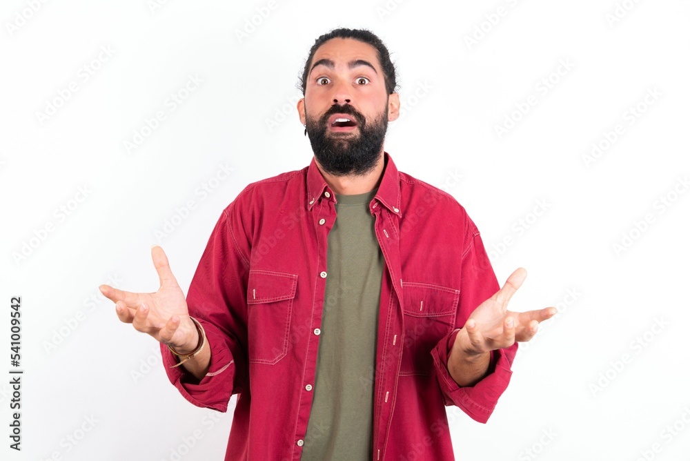 Frustrated Caucasian man with beard wearing red shirt over white background feels puzzled and hesitant, shrugs shoulders in bewilderment, keeps mouth widely opened, doesn't know what to do. Frustrated Caucasian man with beard wearing red shirt over white background feels puzzled and hesitant, shrugs shoulders in bewilderment, keeps mouth widely opened, doesn't know what to do.