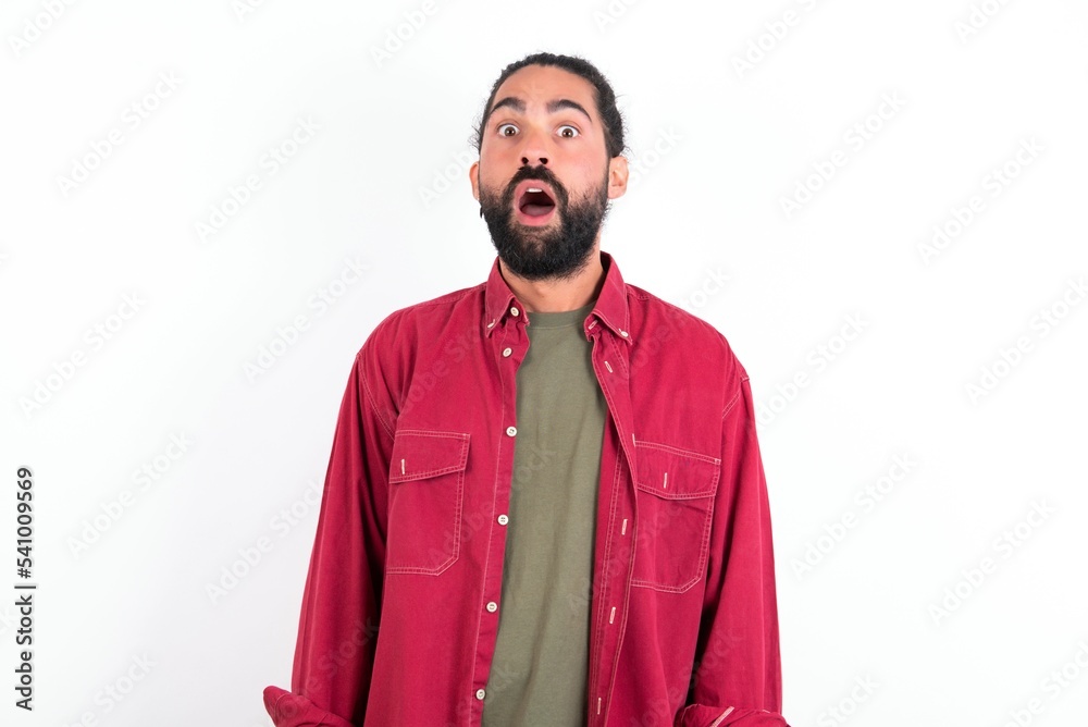 Obraz premium Oh my God. Surprised Caucasian man with beard wearing red shirt over white background stares at camera with shocked expression exclaims with unexpectedness,