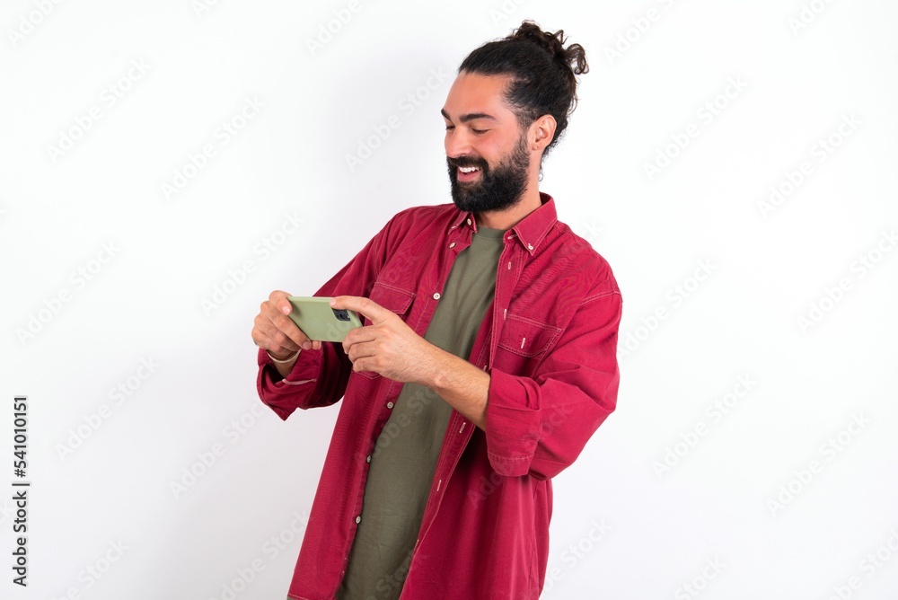 Caucasian man with beard wearing red shirt over white background holding in hands cell playing video games or chatting Caucasian man with beard wearing red shirt over white background holding in hands cell playing video games or chatting