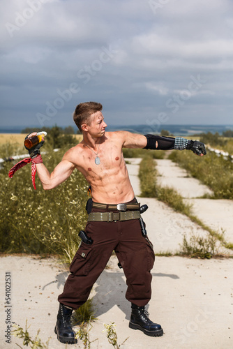 male vandal with dirty naked torso throws Molotov cocktail, against backdrop of an overgrown abandoned road