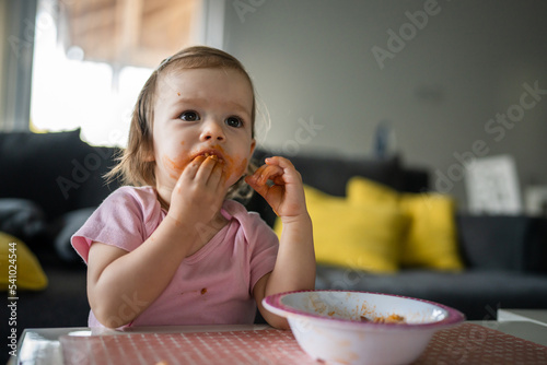 one girl small caucasian toddler female child daughter eating alone at the table at home while watching tv making mess on her face dirty sauce childhood growing up development concept copy space