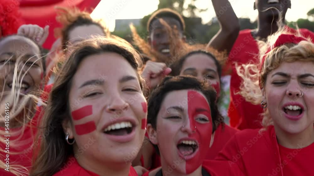 Multiethnic friends football supporter fans streamin a video while watching soccer match at stadium - People with red t-shirts having excited fun on sport world championship event
