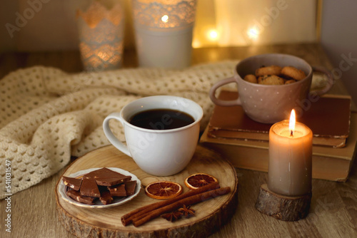 Bowl of cookies, cup of tea or coffee, chocolate, spices, knitted blanket, books, glasses and candle on the table. Cozy hygge atmosphere at home. Selective focus.