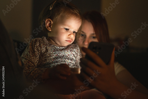 Small caucasian child little girl toddler mother and daughter sitting in dark room at night at home on sofa bed watching cartoons or movies on mobile phone real people family growing up copy space