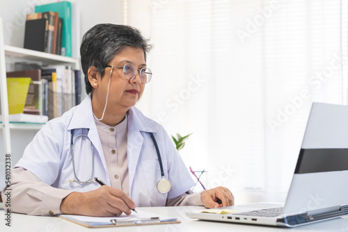 Smile senior asian physician, general woman doctor wearing glasses and white uniform, mature female with stethoscope, working on laptop computer, sitting in medical office. Healthcare and medical.