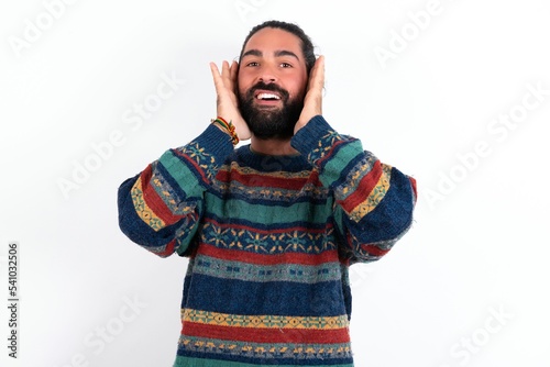 Happy Caucasian man with beard wearing sweater over white background touches both cheeks gently, has tender smile, shows white teeth, gazes positively straightly at camera,