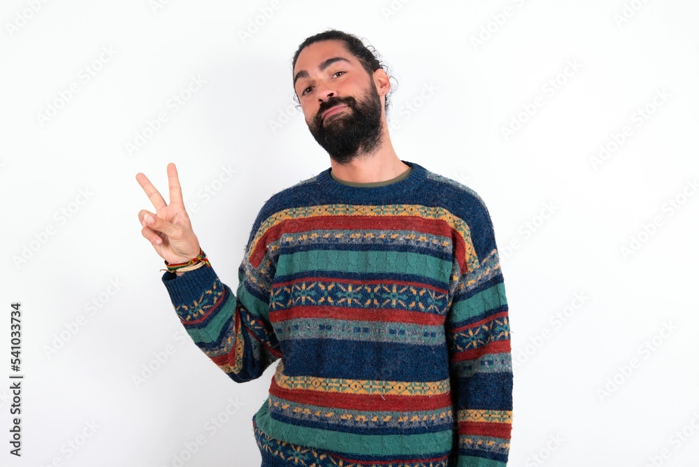 Naklejka premium Caucasian man with beard wearing sweater over white background makes peace gesture keeps lips folded shows v sign. Body language concept