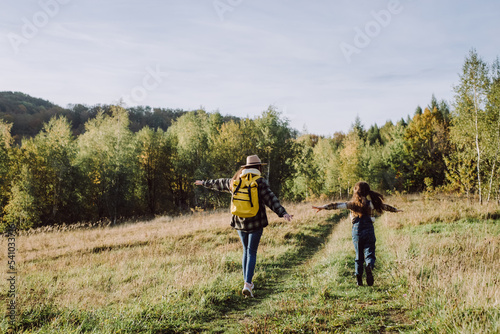 Funny young caucasian mother in hat and playful little daughter child are run grass, playing airplane pilot on background sky and calm fall forest. Traveler family at sunset in beautiful autumn park