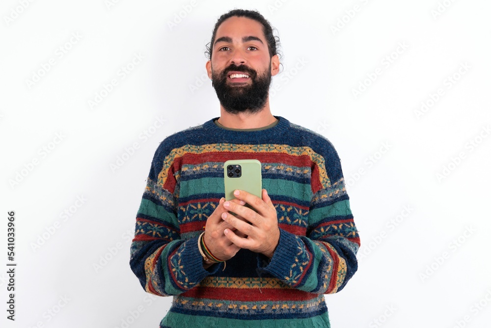 Portrait of serious confident Caucasian man with beard wearing sweater over white background holding phone in two hands Portrait of serious confident Caucasian man with beard wearing sweater over white background holding phone in two hands