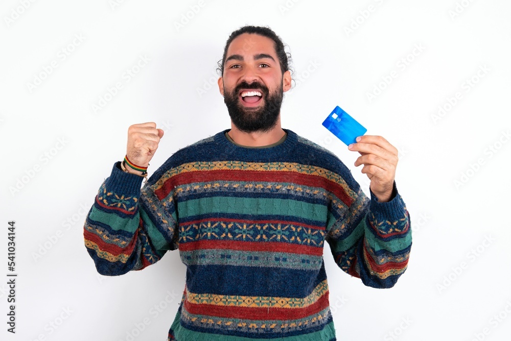 Photo of lucky impressed Caucasian man with beard wearing sweater over white background arm fist holding credit card. Celebrated Photo of lucky impressed Caucasian man with beard wearing sweater over white background arm fist holding credit card. Celebrated