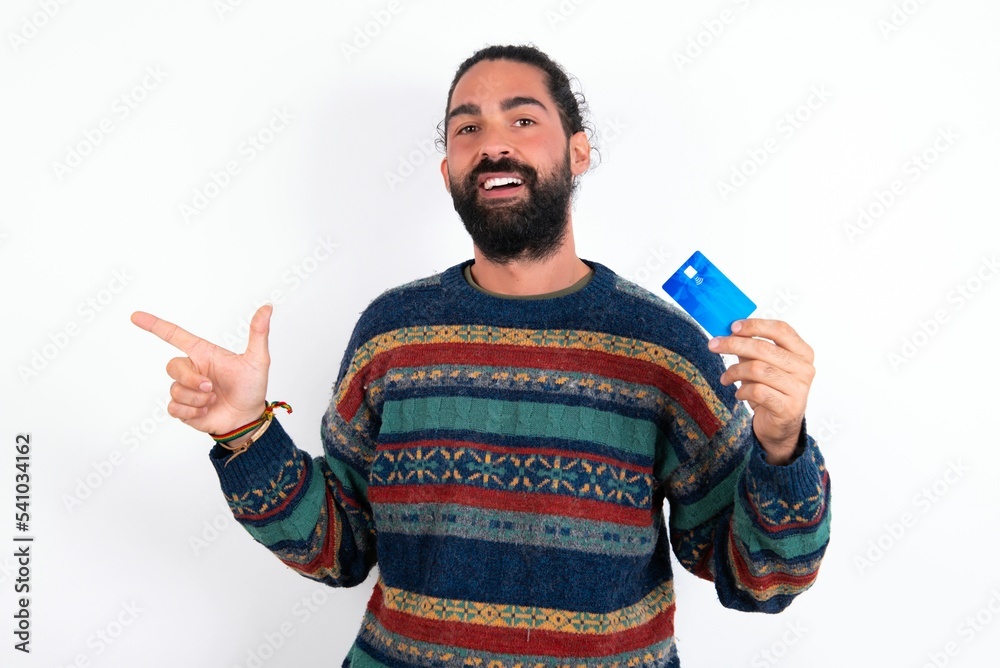 Curious smiling Caucasian man with beard wearing sweater over white background showing plastic bank showing finger copyspace Curious smiling Caucasian man with beard wearing sweater over white background showing plastic bank showing finger copyspace