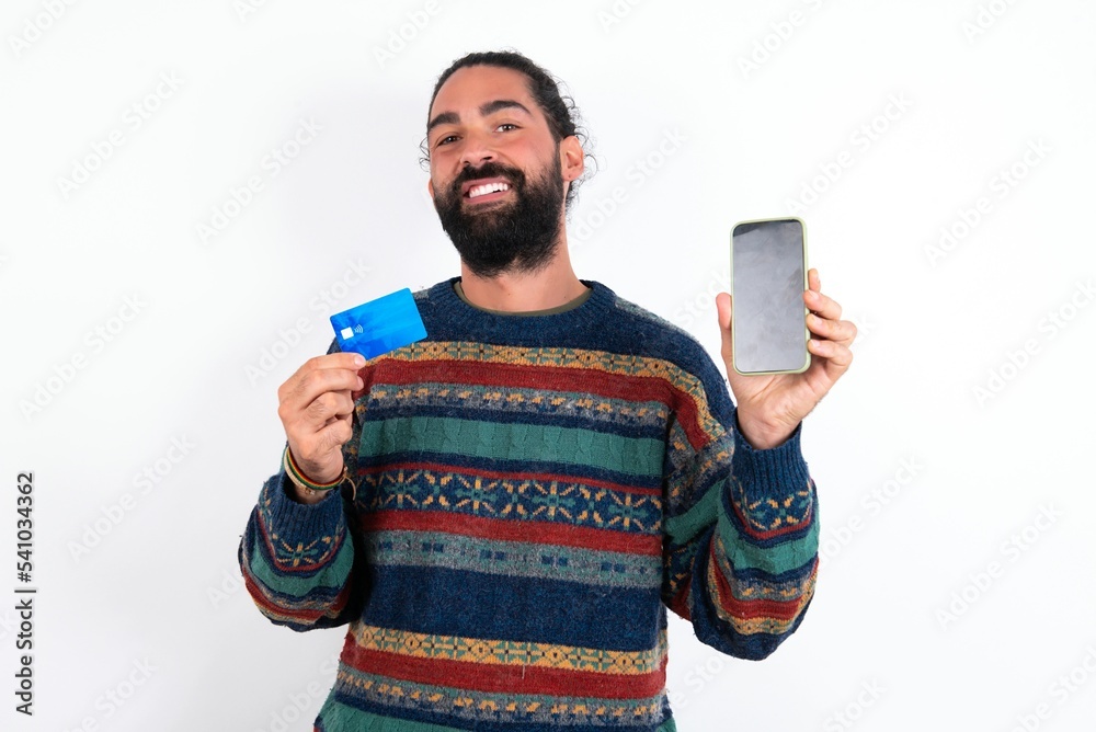 Caucasian man with beard wearing sweater over white background wears stereo headphones on ears holds modern mobile phone and credit card Caucasian man with beard wearing sweater over white background wears stereo headphones on ears holds modern mobile phone and credit card