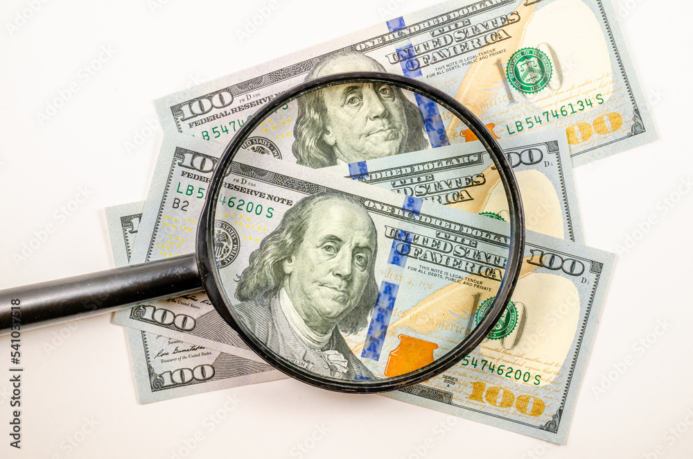 One hundred US dollars under a magnifying glass . One hundred US dollars under a magnifying glass .