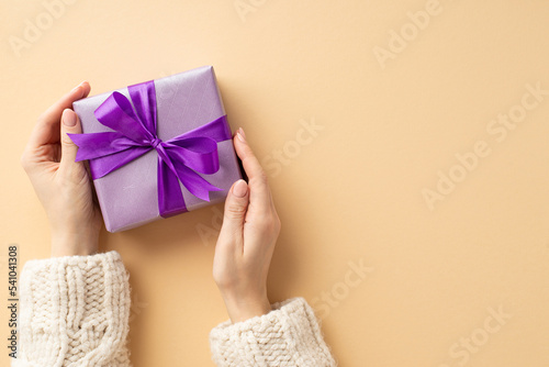 Fototapeta Naklejka Na Ścianę i Meble -  Christmas Day concept. First person top view photo of female hands in white knitted sweater holding lilac giftbox with violet ribbon bow on isolated pastel beige background with copyspace