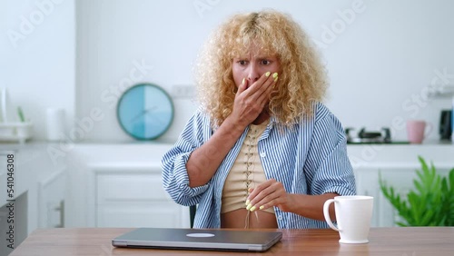 Young anxious frustrated Caucasian curly-haired blonde woman, experiencing fright, fear and sense of horror, feeling shocked by what she has seen, she closes the lid of the laptop computer