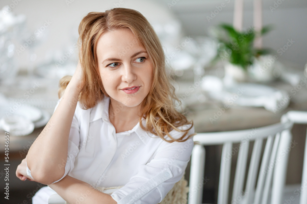 Beautiful smiling young woman with long blonde hair sitting at table in light white  room