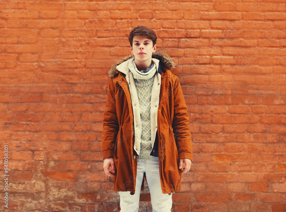 Portrait of stylish young man wearing brown jacket, knitted sweater in the city on textured brick wall background