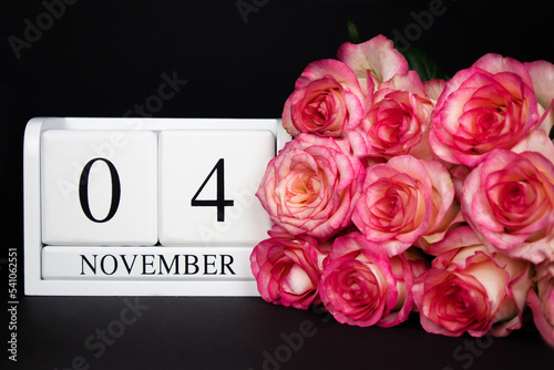 November 4 wooden calendar, white on a black background, pink roses lie nearby. Postcard with copy space. The concept of a holiday, congratulation, invitation, party, announcement, vacation,promotion. © Anna