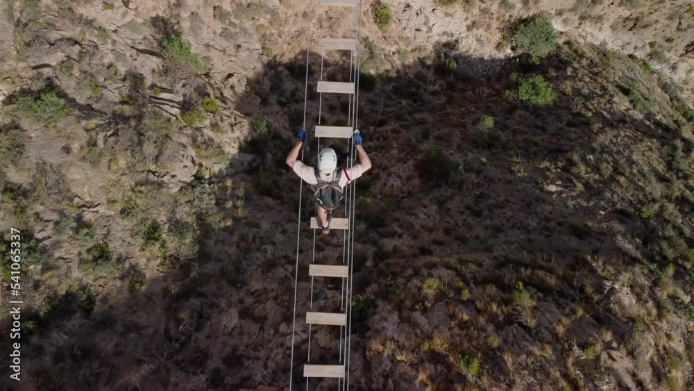 Man overcomes his fears going through a suspension bridge of a via ferrata, bird's eye view, man equipped with harness, helmet and ropes. Man overcomes his fears going through a suspension bridge of a via ferrata, bird's eye view, man equipped with harness, helmet and ropes.