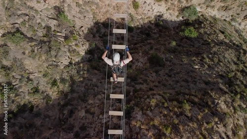 Man overcomes his fears going through a suspension bridge of a via ferrata, bird's eye view, man equipped with harness, helmet and ropes.