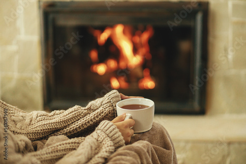 Hands in cozy sweater holding cup of warm tea on background of burning fireplace close up, autumn hygge. Heating house with wood burning stove. Relaxing and warming up at rustic fireplace photo