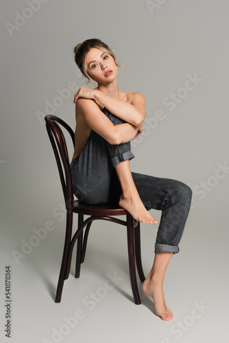pretty barefoot and half dressed woman in jeans sitting on wooden chair and looking at camera on grey background