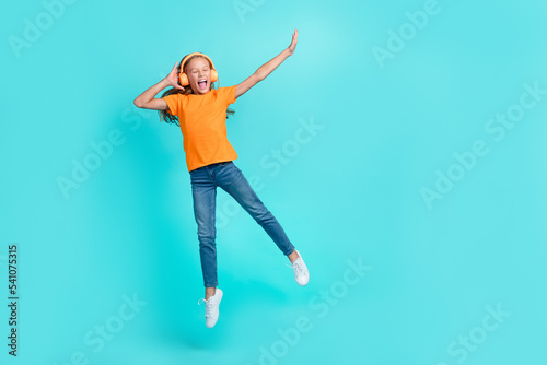 Full length photo of cheerful positive little child dressed orange t-shirt earphones jumping high empty space isolated teal color background