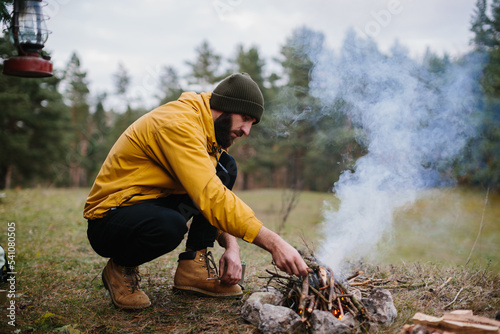 Survival in the wild. A bearded man lights a fire near a makeshift shelter made of pine branches.