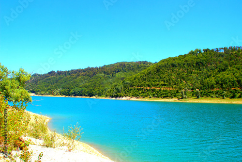 Clear view of Gerosa Lake with its slightly rippled pristine glowing azure waters sitting between the greenery-topped shores and Marche hills under a cloudless blue sky on a fine summer day