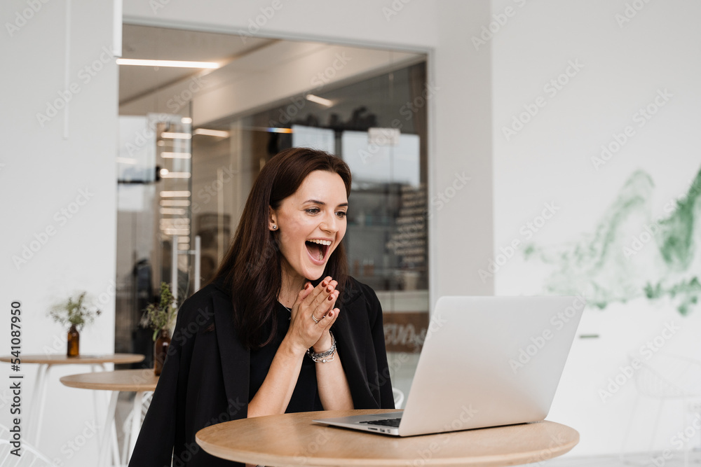 Happy girl rejoices in winning and sitting in front of laptop in white cafe. Cheerful surprised woman smile and looking at amount of income earnings salary and rejoices. Happy girl rejoices in winning and sitting in front of laptop in white cafe. Cheerful surprised woman smile and looking at amount of income earnings salary and rejoices.