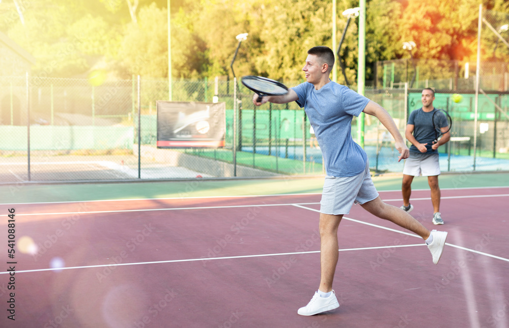 Young teenage tennis player training on court with male partner. Boy using racket to hit ball.