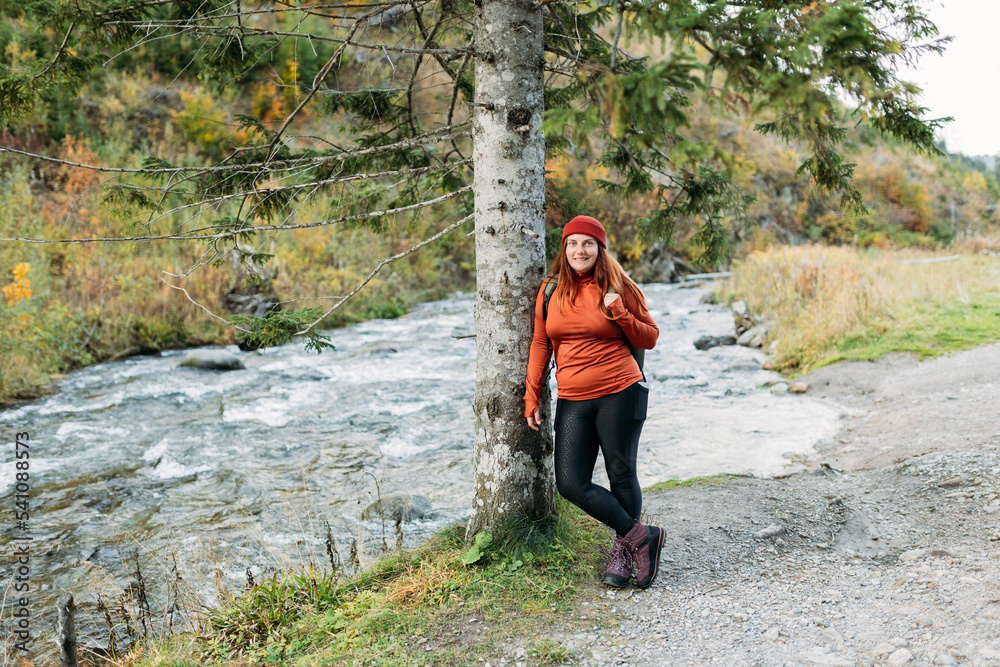 Happy redhead Woman in red hat hiking in woods. Adventure women standing near the mountain river. Freedom and active lifestyle concept. Amazing chill moment