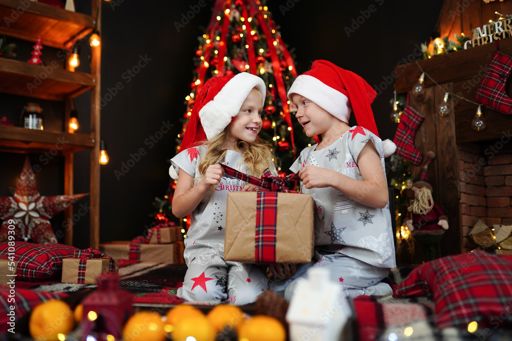 Children in pajamas in Santa's hats opening gift box while sitting near fireplace and Christmas tree Children in pajamas in Santa's hats opening gift box while sitting near fireplace and Christmas tree