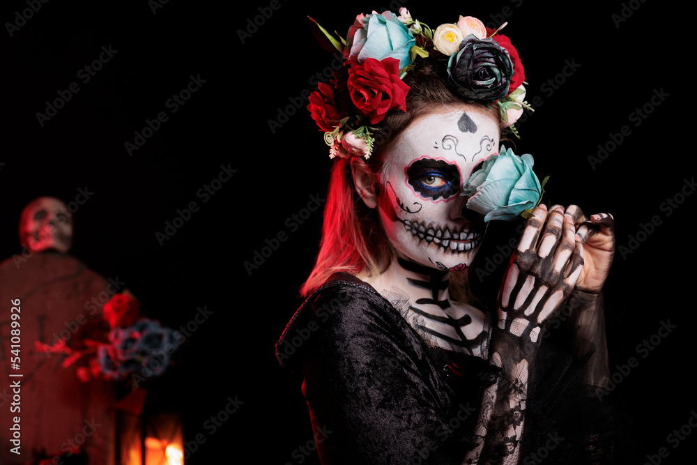 Day of the dead skull model with body art celebrating mexican holiday tradition with santa muerte costume. Posing with roses and festival make up, flowers crown on studio background.