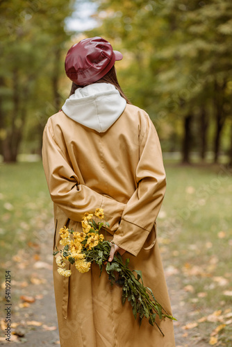 Unrecognizable stylish woman in trench and cap walking with bouquet of yellow flowers behind back in autumn park 