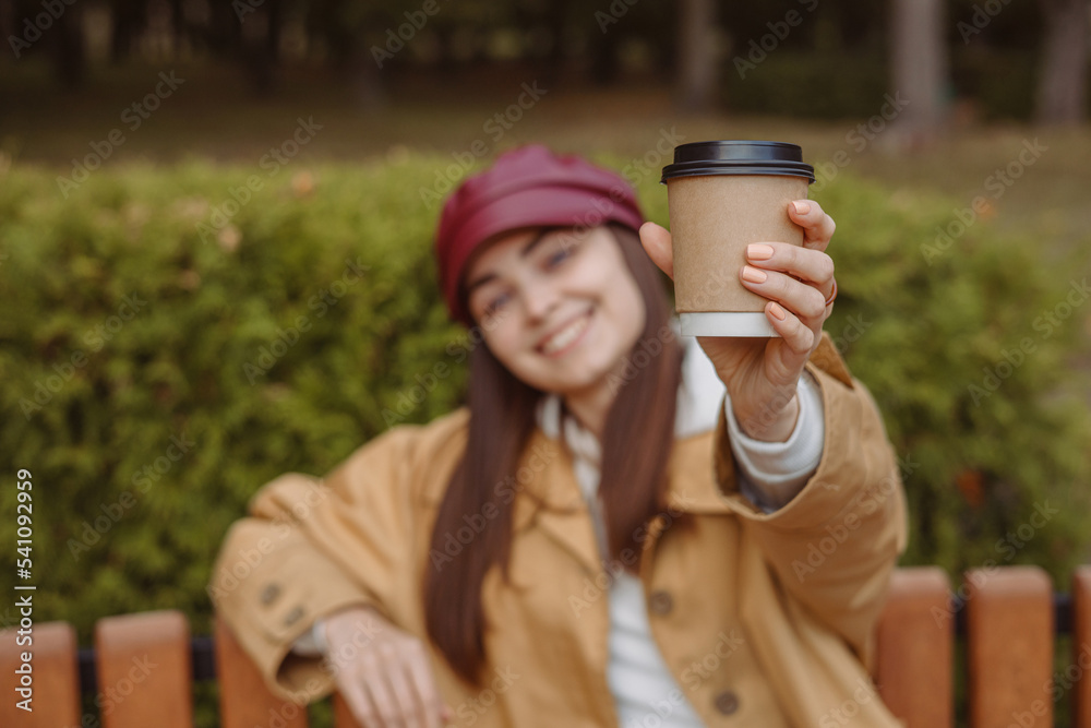 Blurred smiling woman showing blank paper cup with takeaway coffee while sitting on bench in park in autumn 