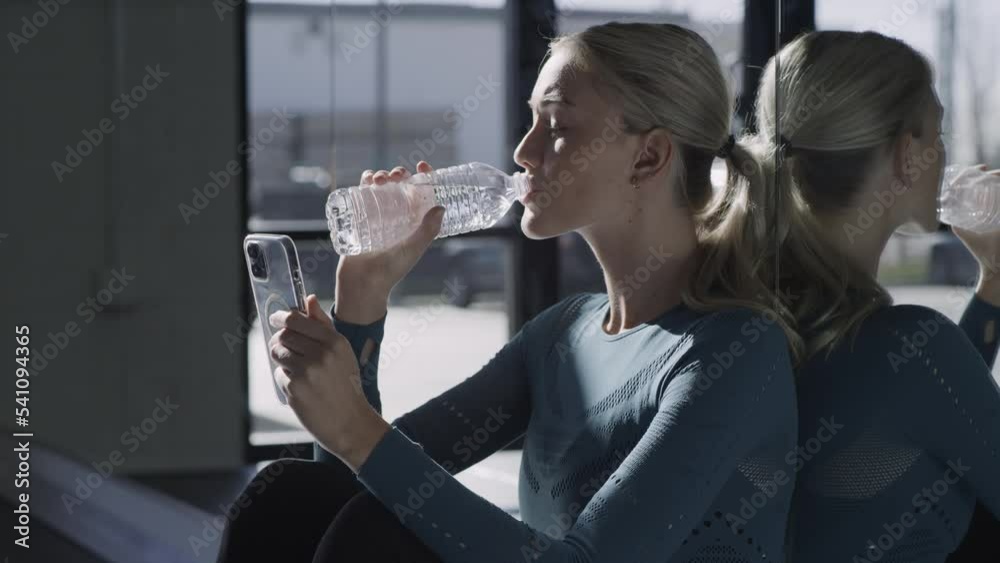 Woman sitting on floor leaning on mirror wall drinking water and checking cell phone / Lehi, Utah, United States Woman sitting on floor leaning on mirror wall drinking water and checking cell phone / Lehi, Utah, United States