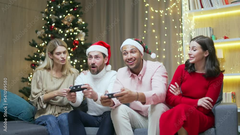 New Year's party group of four diverse friends having fun relaxing and celebrating on Christmas holidays guests sitting on sofa men and women playing video games on joystick consoles Conflict interest New Year's party group of four diverse friends having fun relaxing and celebrating on Christmas holidays guests sitting on sofa men and women playing video games on joystick consoles Conflict interest