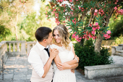 Groom hugs bride from behind by the shoulders under a blooming oleander